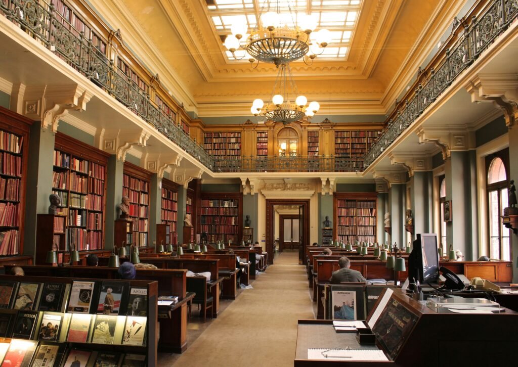 Readers at wooden desks beneath bookshelves and chandeliers in London’s National Art Library.